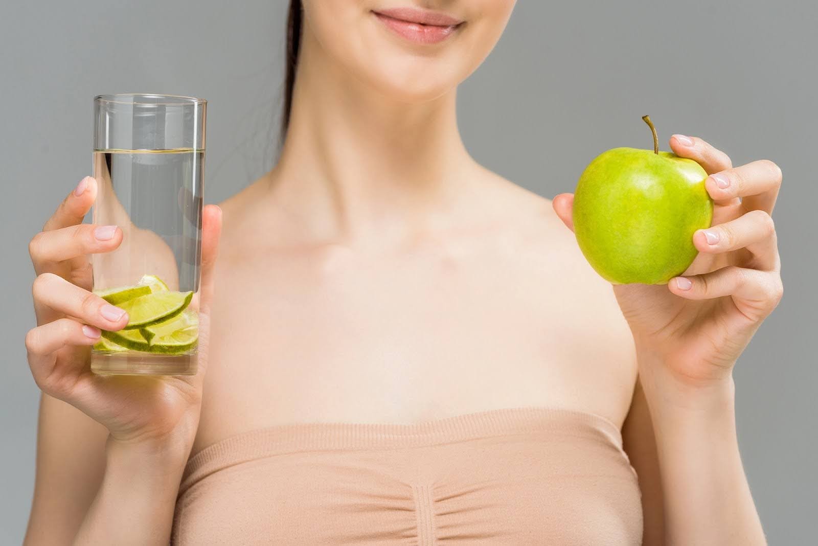 A person holding a glass of water with lime slices in one hand and a green apple in the other