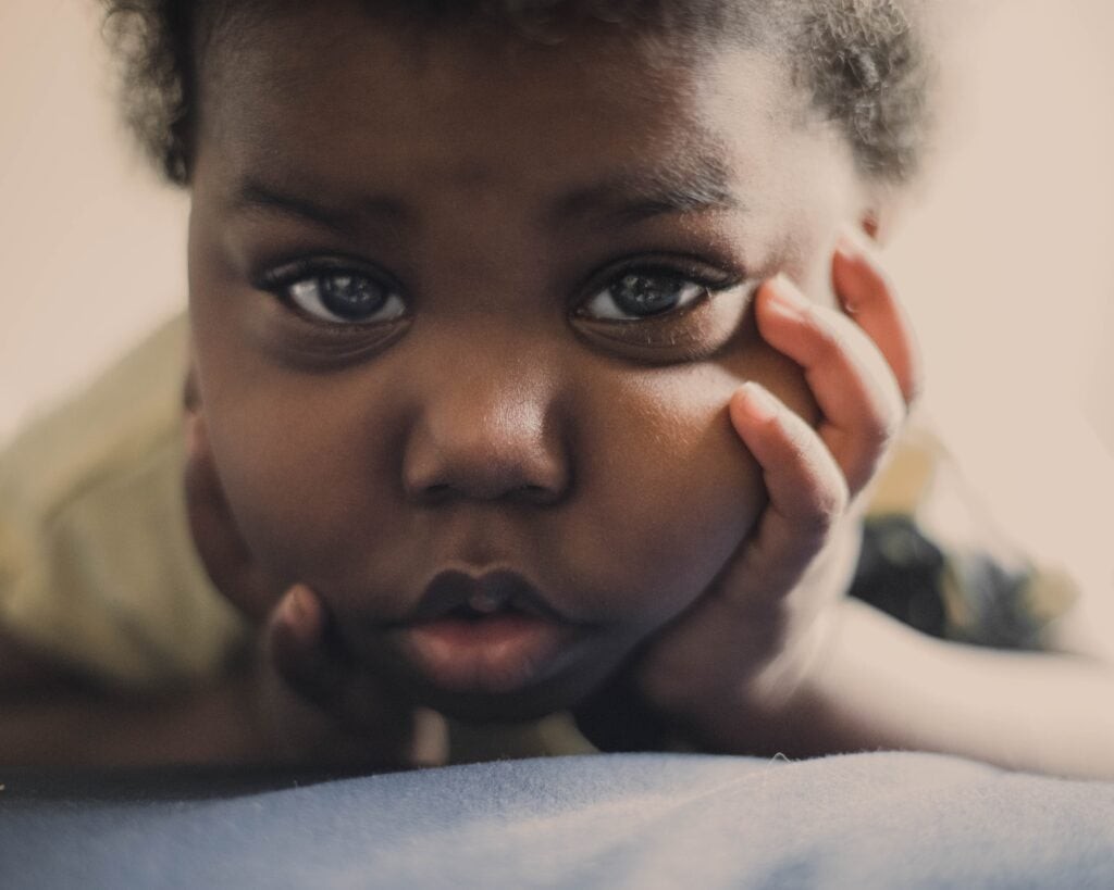 A close-up portrait of a young child resting their chin on their hand, looking directly at the camera