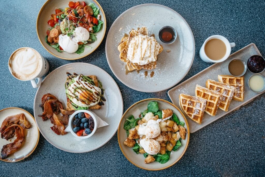 A top-down view of a table spread with various breakfast foods, including waffles, poached eggs, bacon, berries, and coffee