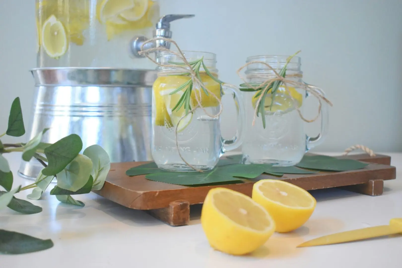 Glass mason jars of lemon water with rosemary on a wooden board with fresh lemons