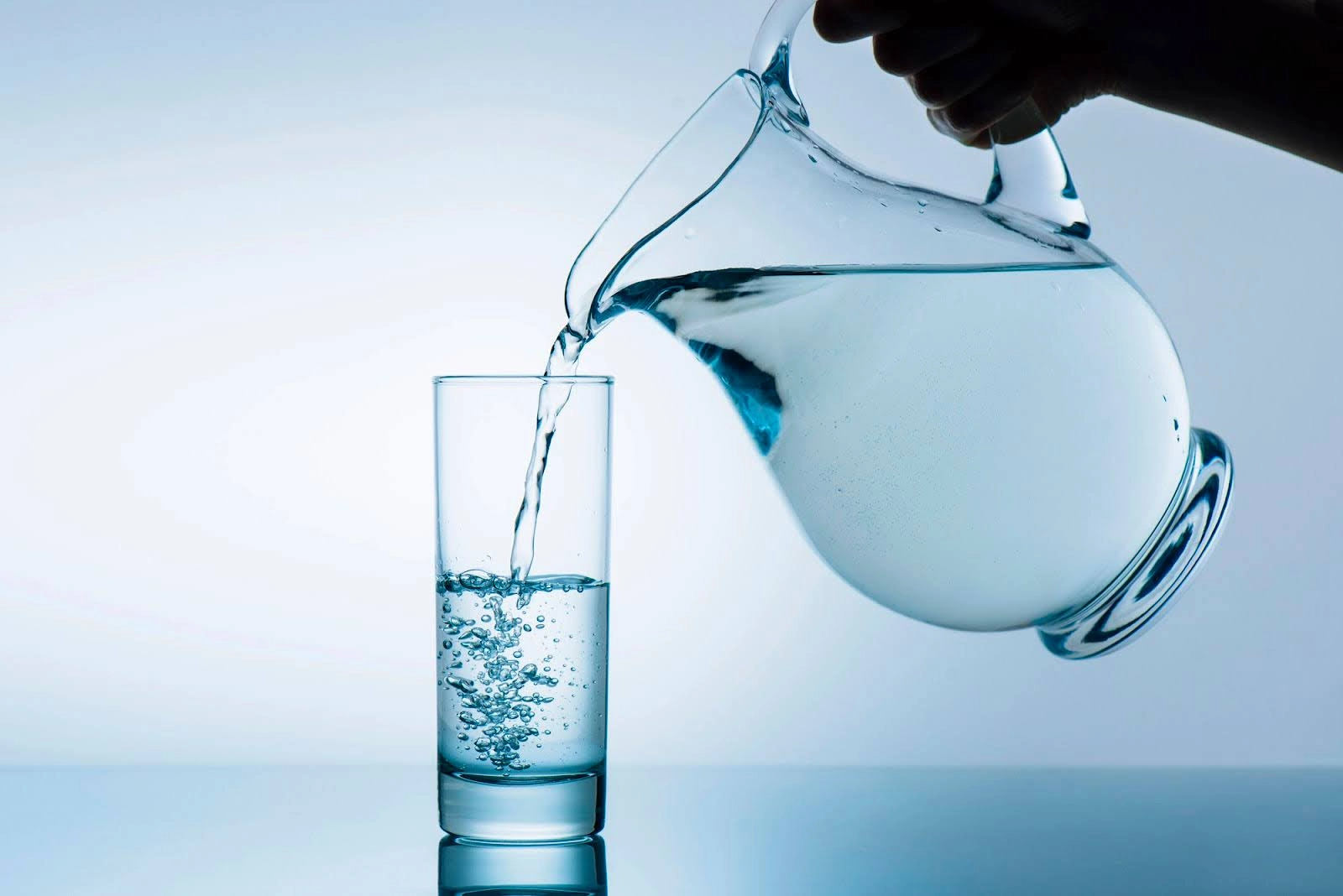Water being poured from a clear pitcher into a tall glass on a reflective surface