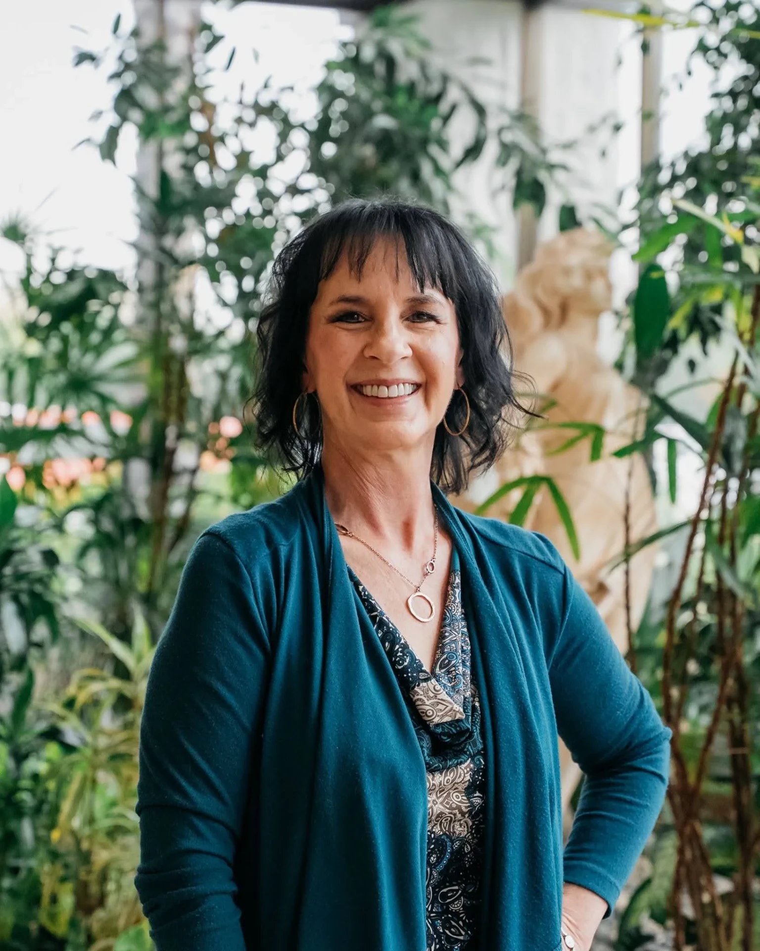 Smiling woman with dark hair in a blue cardigan standing in front of lush green plants