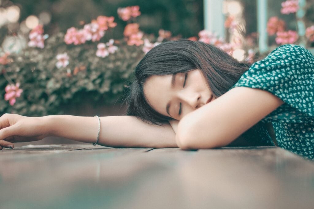 A woman sleeping with her head resting on her arms on a wooden surface, with flowers in the background