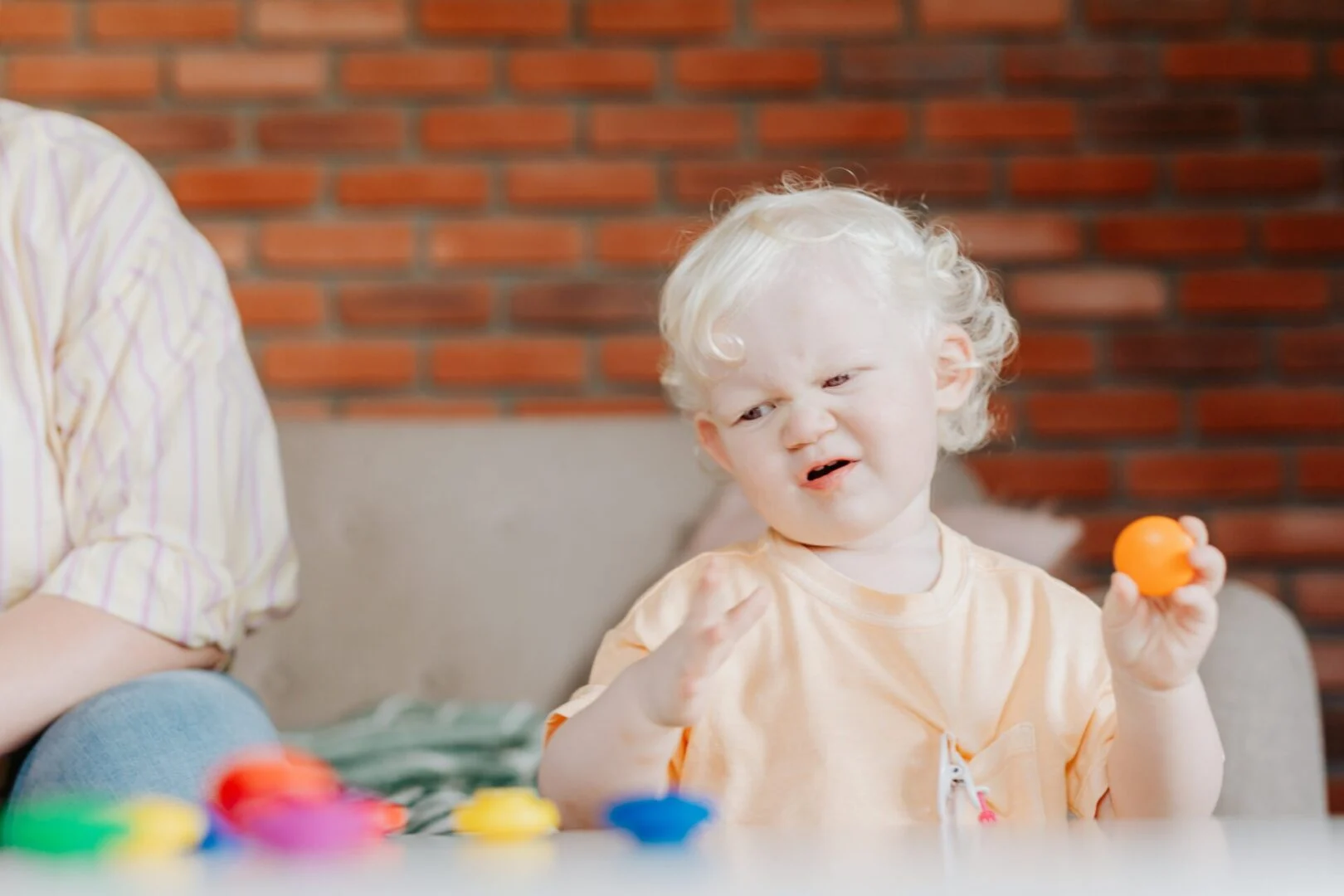 Toddler with curly blonde hair making a fussy expression while holding a small orange ball